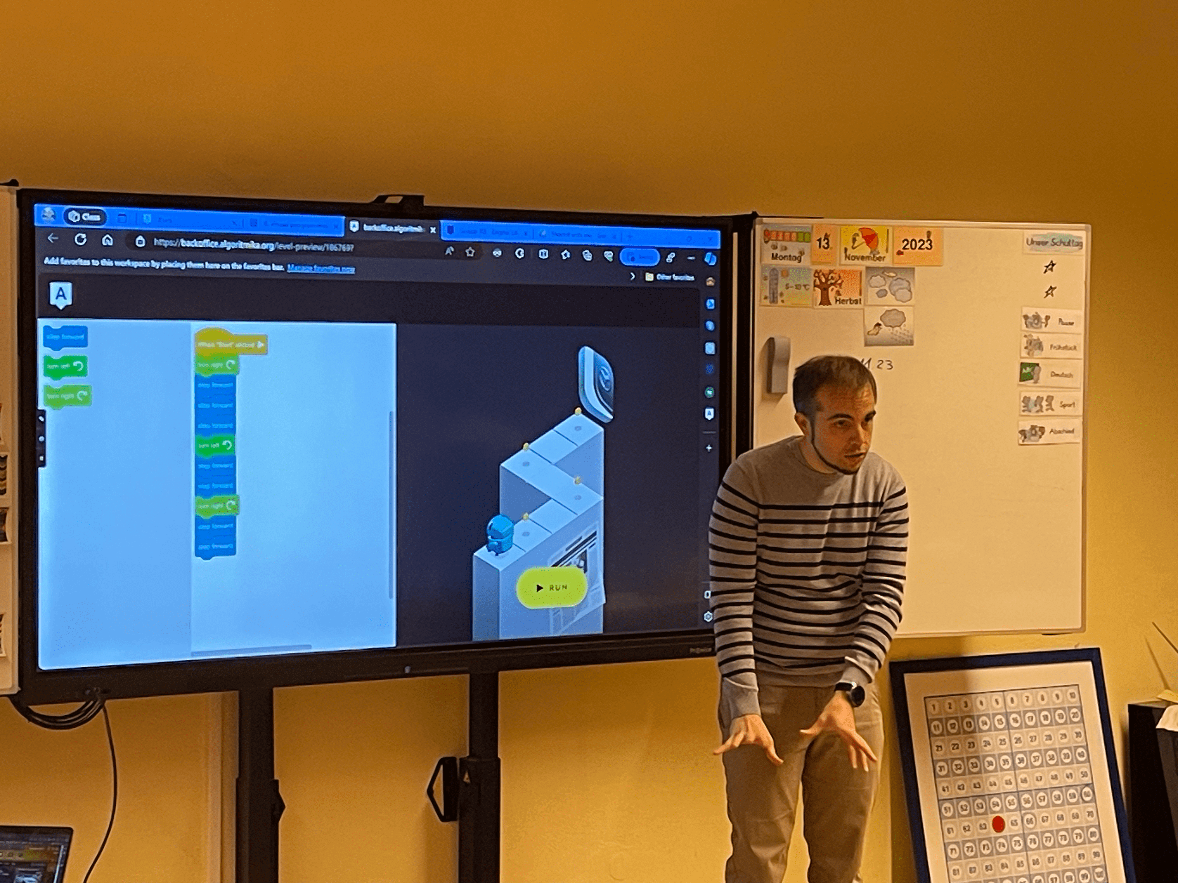 Elias, a teacher, stands in front of a large monitor displaying a block-based programming interface, actively engaging with the class. The screen shows a coding exercise, while Elias gestures to explain the concept. A calendar and educational posters adorn the classroom wall. - Algorithmics School Dusseldorf