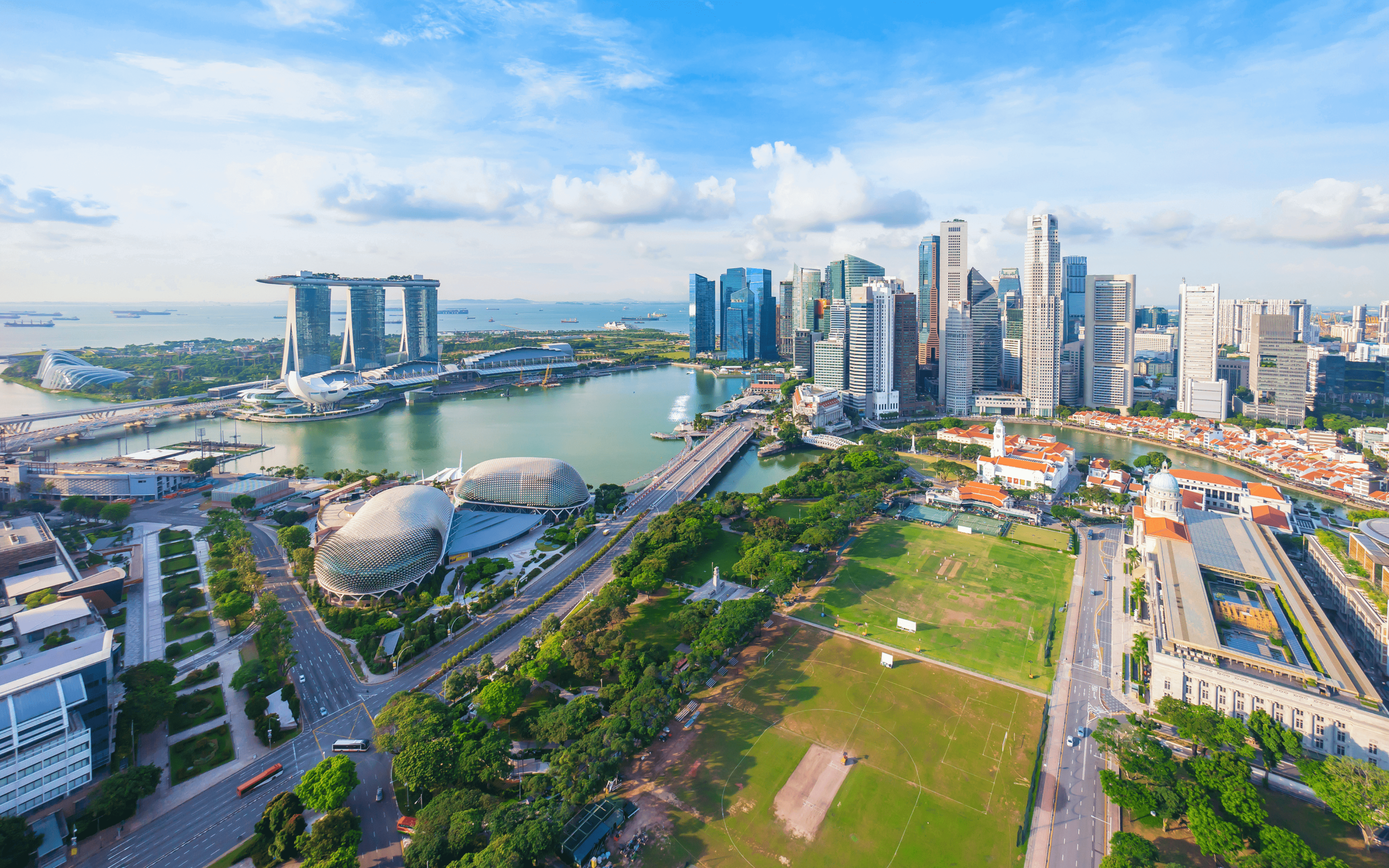 Aerial view of Singapore's iconic skyline including the Marina Bay Sands, the Esplanade Theaters, and the verdant greenery of the Padang. The city is a tapestry of modern architecture, lush parks, and bustling waterways.