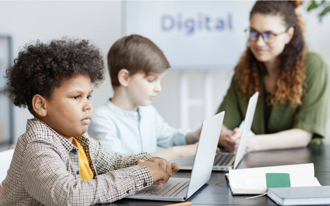 Two children focused on their laptops in a modern classroom setting with a teacher assisting in the background, the word 'Digital' displayed on a screen indicating a technology-driven education theme.