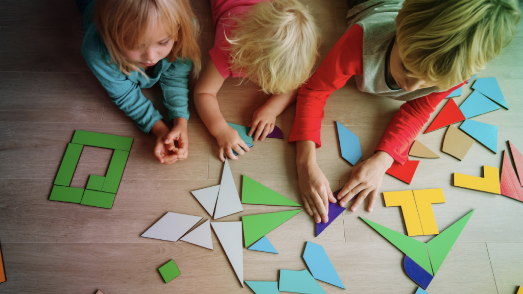 Three young children collaboratively playing with colorful geometric shape blocks on a wooden floor, arranging them into patterns and structures.